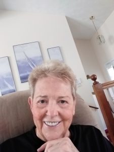 Kay Cavanaugh, smiling, with short grey hair, sitting in her arm chair in the living room.