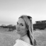 Black-and-white image of Jennifer House, hair loose and flowing, sunglasses propped on her head, off-the shoulder white blouse, looking over her shoulder as she walks down a beach.