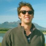 Image of JJ Graham, outside, smiling, his hair caught in the wind, wearing dark sunglasses and a fleece pullover.