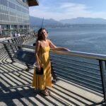 Image of Suzette Blom, wearing a yellow sun dress and sandals, posing outside by a walkway, over looking a view of water and mountains.